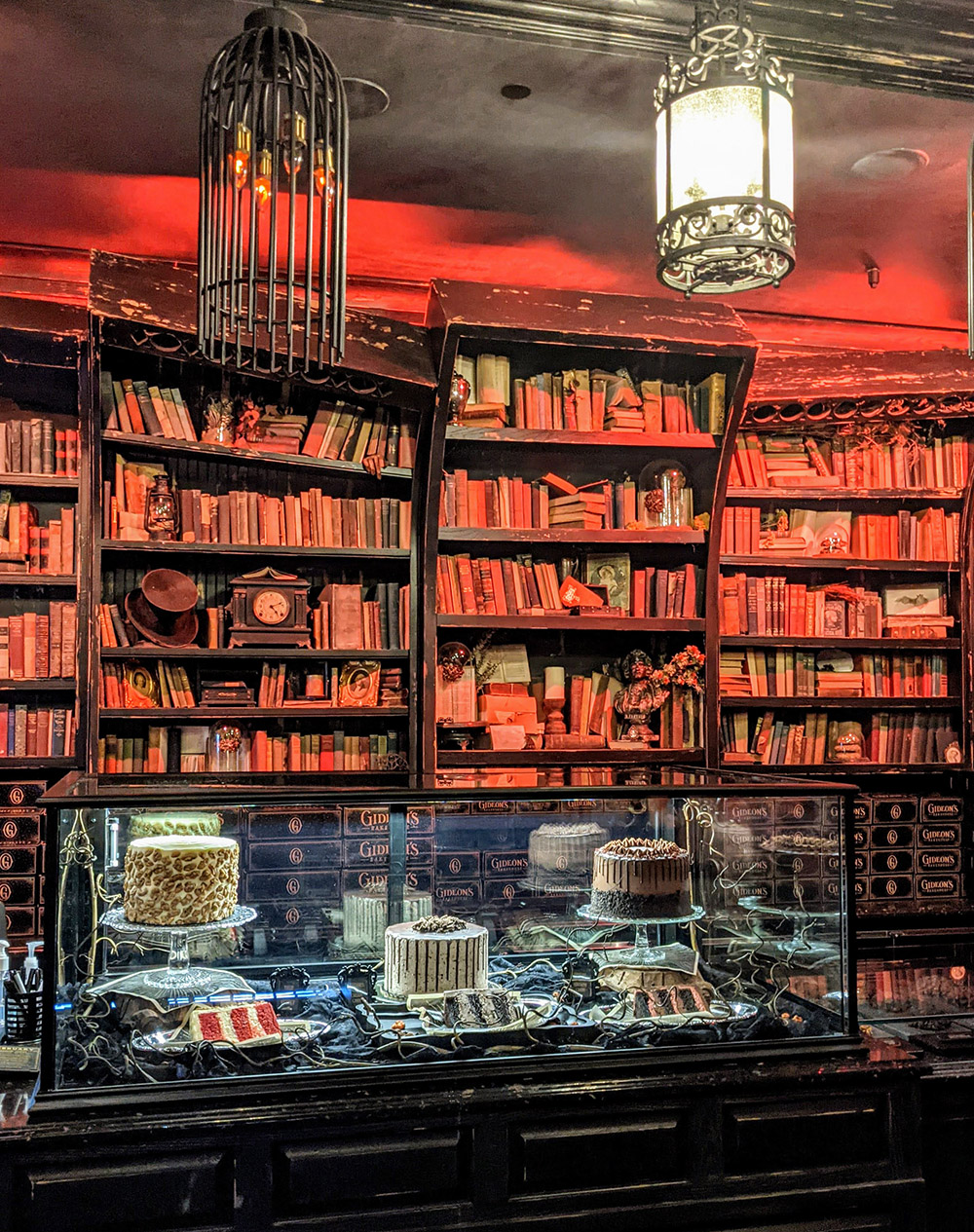 What it's like inside Gideon's Bakehouse in Disney Springs: old books line the walls from floor to ceiling, there's a reddish tint to the lights, and desserts are displayed under glass like artifacts