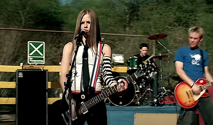 Avril Lavigne wearing a black tie, tank top and striped arm warmer while strumming a guitar outside