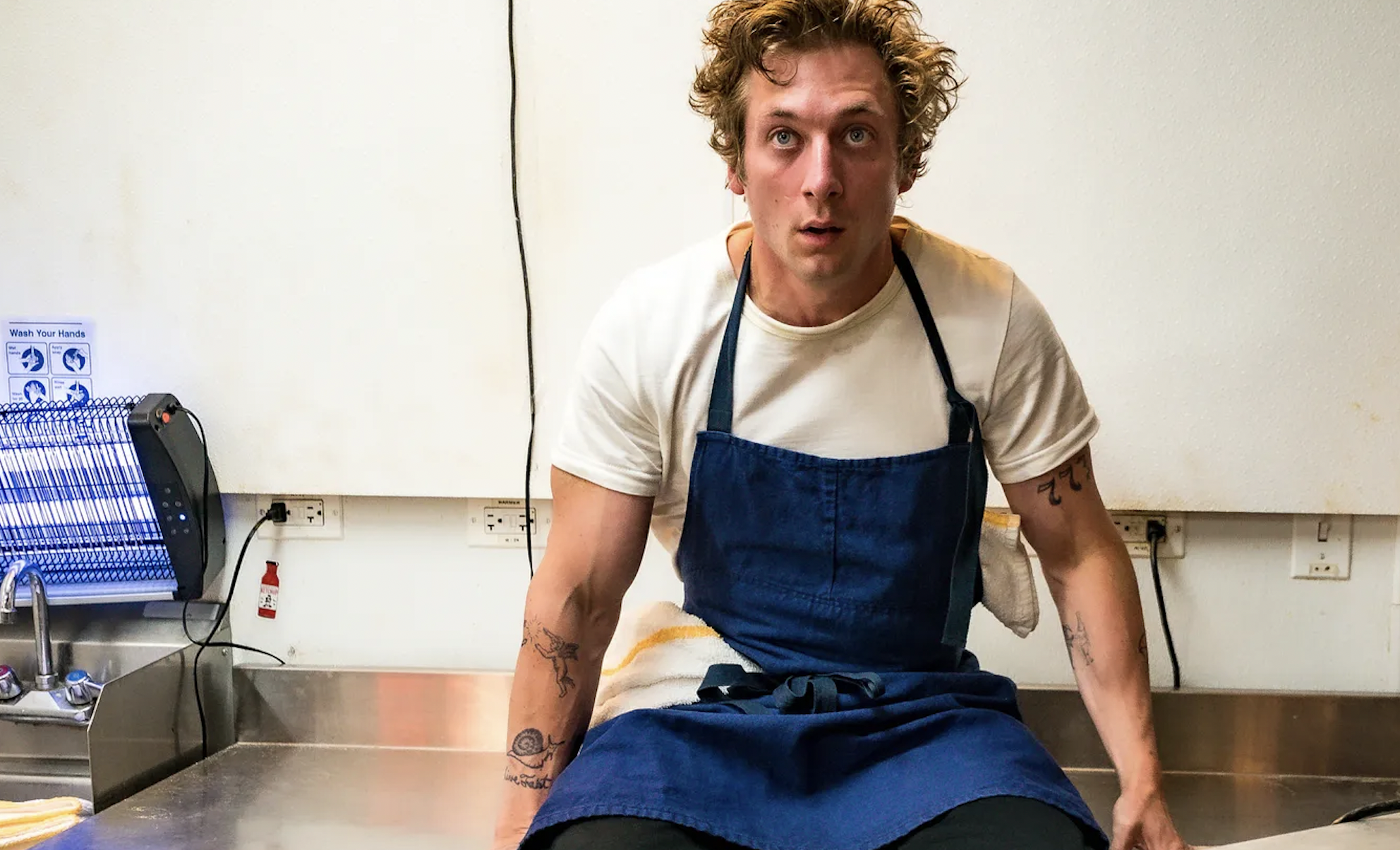 Carmy in 'The Bear' wearing a white shirt, blue apron and dark pants